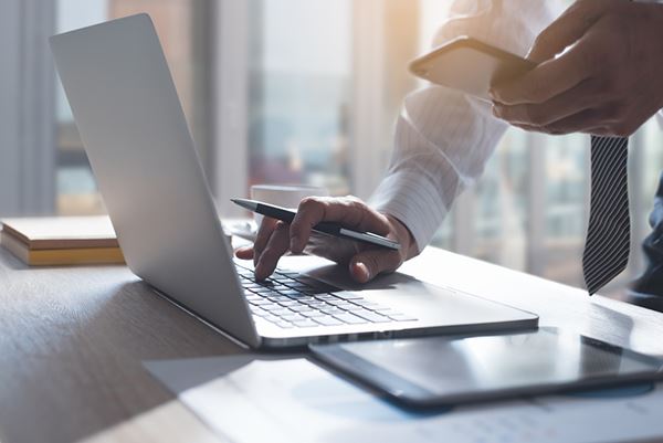 A businessman types on his laptop while holding his phone.