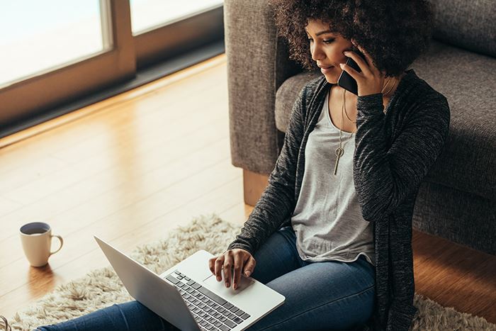 A woman sitting on the floor on her phone and a her laptop.